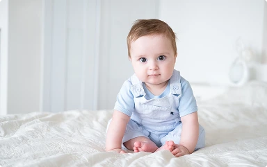 baby-boy-6-months-old-blu-clothes-smiling-sitting-white-bed-home 1